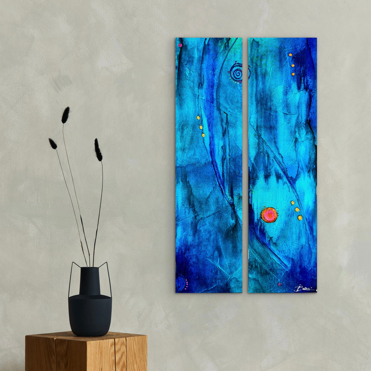 A two-panel abstract wall art print with vivid blue and teal brushstrokes and small red and gold accents, displayed on a textured light gray wall above a wooden block with a black vase.