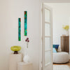 Two tall, narrow abstract wall art panels in teal, green, orange, and blue hang on a white wall in a bright interior hallway. A white door is open to reveal a softly lit living room with a round pouf, blue and beige pillows, a wooden cabinet, and a vase with yellow flowers.