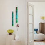 Two tall, narrow abstract wall art panels in teal, green, orange, and blue hang on a white wall in a bright interior hallway. A white door is open to reveal a softly lit living room with a round pouf, blue and beige pillows, a wooden cabinet, and a vase with yellow flowers.