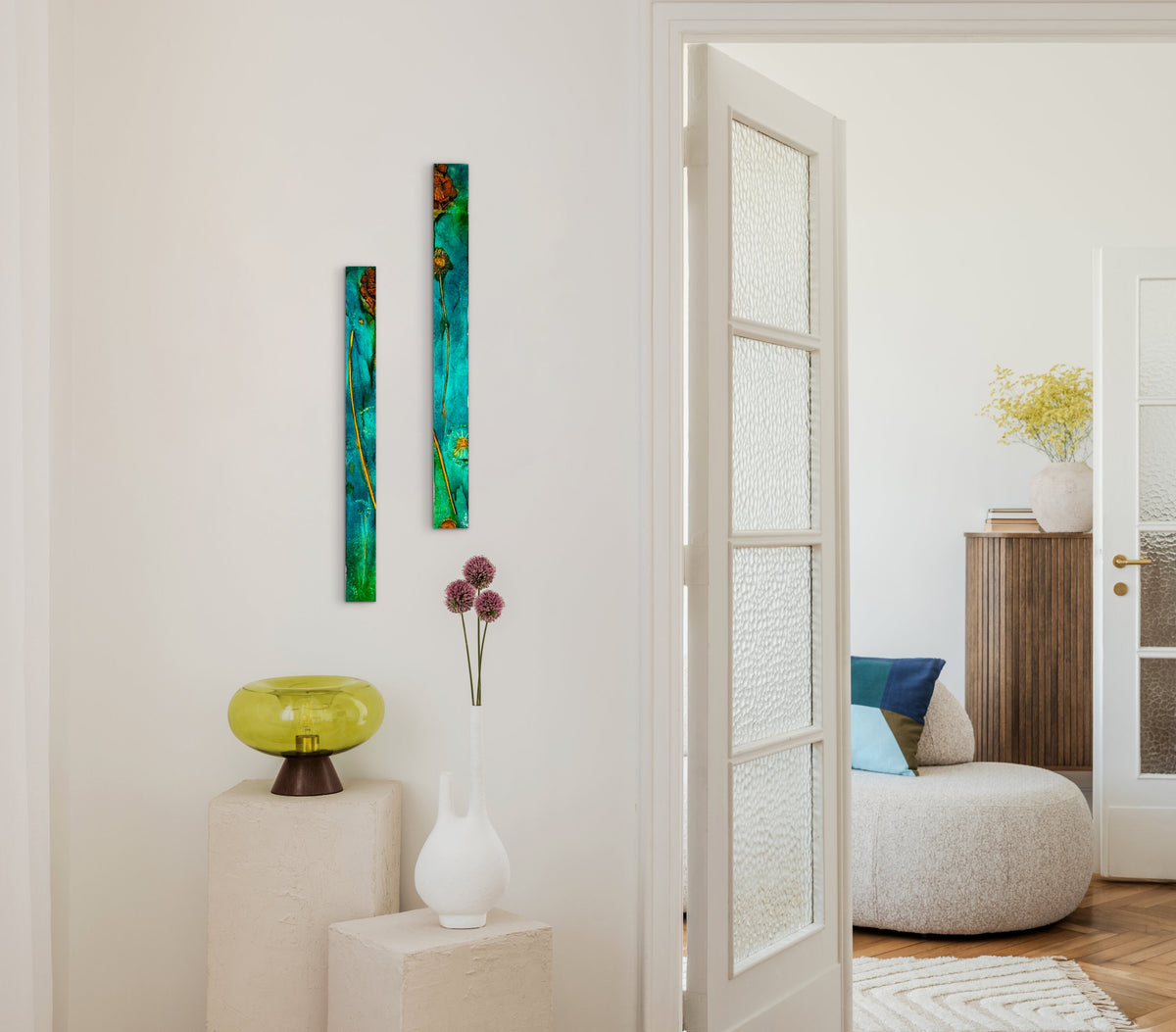 Two tall, narrow abstract wall art panels in teal, green, orange, and blue hang on a white wall in a bright interior hallway. A white door is open to reveal a softly lit living room with a round pouf, blue and beige pillows, a wooden cabinet, and a vase with yellow flowers.