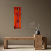 A vertical three-panel canvas wall art print featuring a vibrant red and orange abstract tree design, displayed on a taupe wall above a rustic wooden bench with a small vase of branches.
