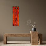 A vertical three-panel canvas wall art print featuring a vibrant red and orange abstract tree design, displayed on a taupe wall above a rustic wooden bench with a small vase of branches.