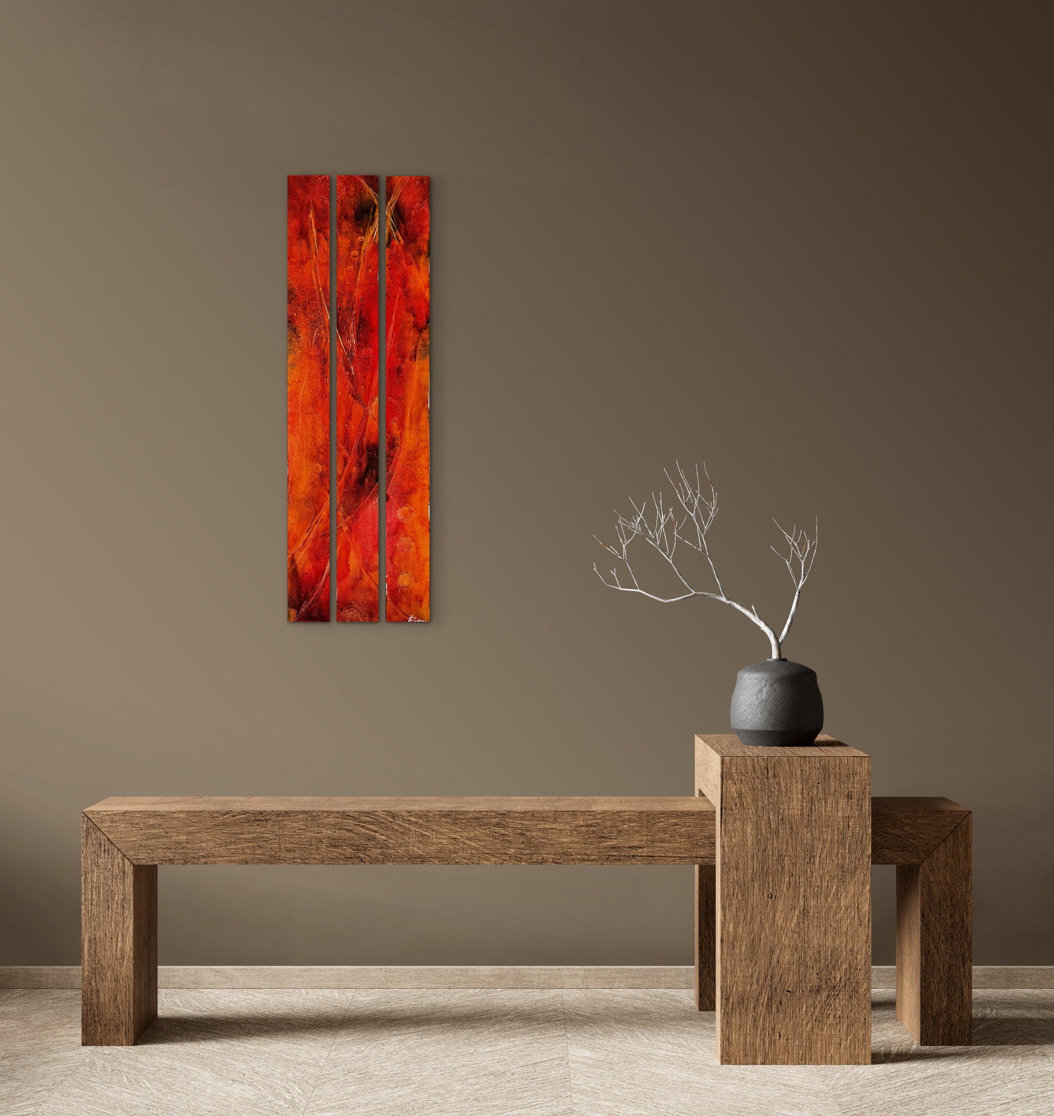 A vertical three-panel canvas wall art print featuring a vibrant red and orange abstract tree design, displayed on a taupe wall above a rustic wooden bench with a small vase of branches.